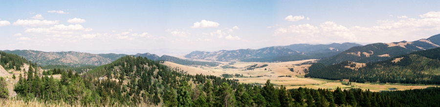 [Several photos stitched together giving a panoramic view of the mountains and valleys. Evergreens cover most of the mountains while the valleys are light brown and have little vegetation.]